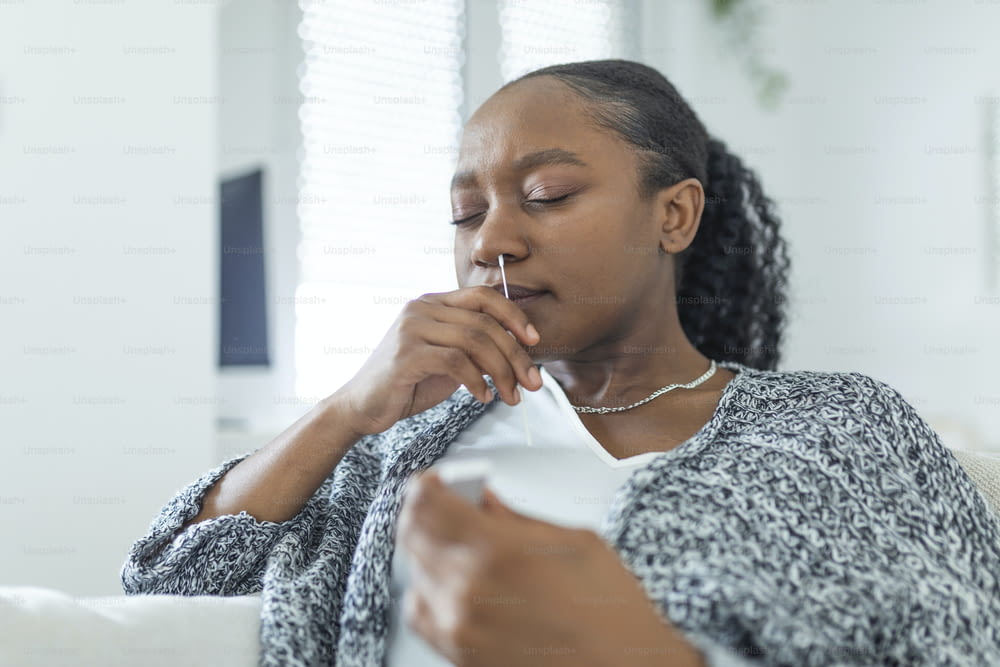 집에서 코로나바이러스 PCR 검사를 하는 동안 면봉을 사용하는 아프리카계 미국인 여성. 코로나바이러스 신속 진단 테스트를 사용하는 여성. COVID-19에 대해 비강 면봉을 사용하는 집에서 젊은 여성.