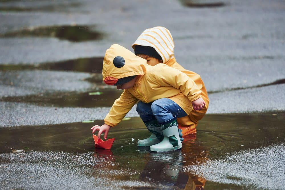 Duas crianças em capas e botas amarelas impermeáveis brincando com barco de papel artesanal ao ar livre após a chuva juntos.