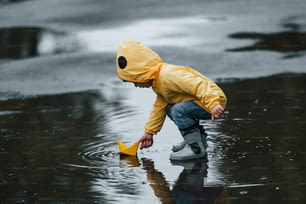 Niño con capa impermeable amarilla y botas jugando con un juguete de barco hecho a mano de papel al aire libre después de la lluvia.