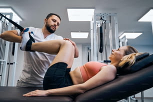 スポーツ傷害をリハビリするための弾性マシンでの若い女性の脚のマッサージと筋力トレーニング。男性理学療法士が理学療法治療で脊椎と背中を治療