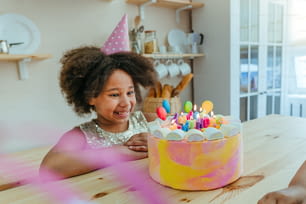Menina feliz olhando para o bolo de aniversário com velas se divertindo durante a festa de aniversário na cozinha. Foco seletivo no rosto da menina.
