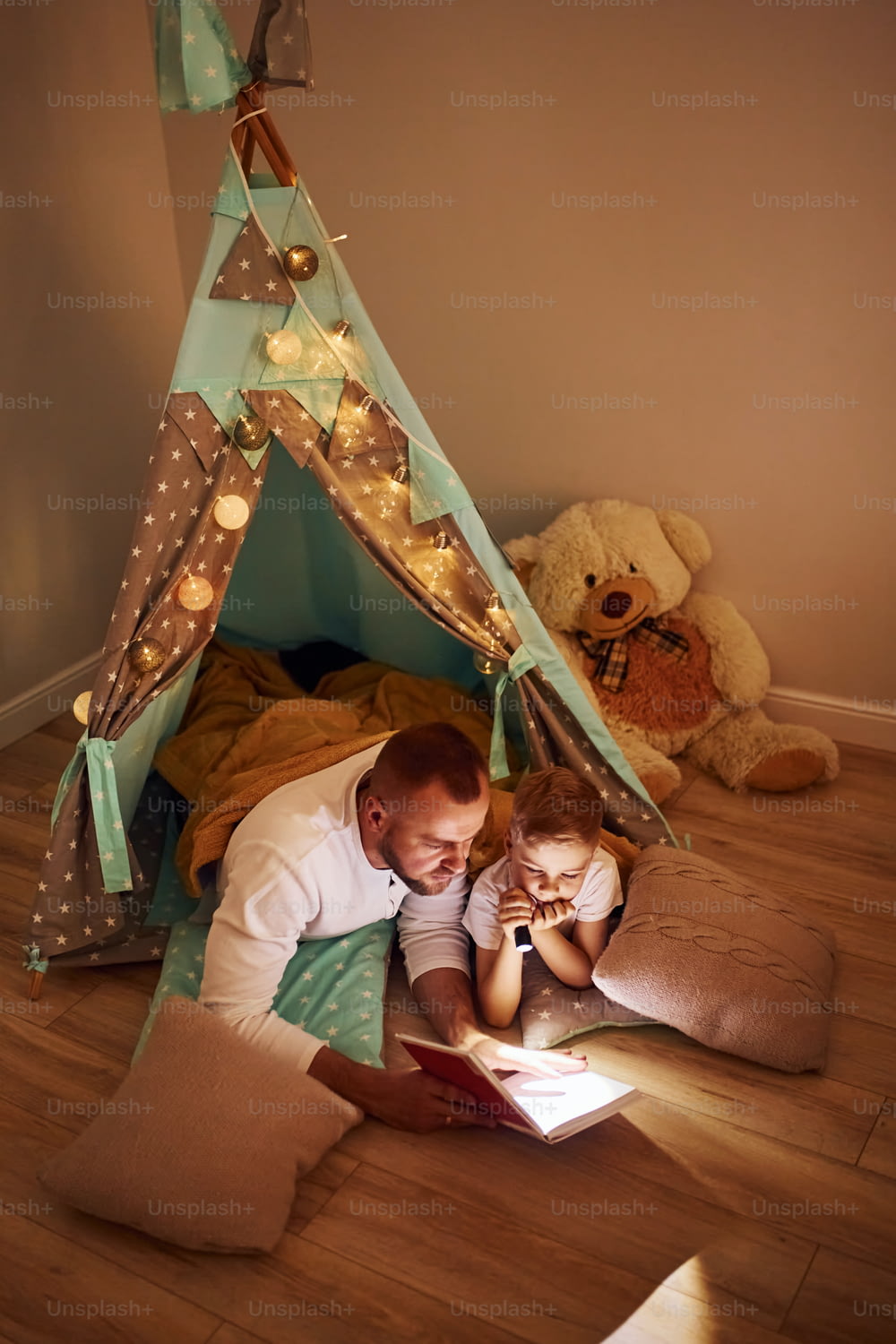 Reading book and using flashlight. Father and his young young spending time together indoors near tent.