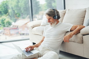 Sits on the ground near sofa with laptop and listens music. Senior stylish modern man with grey hair and beard indoors.