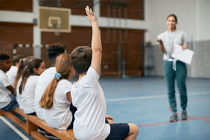 体育館で体育の授業を受けながら、腕を上げて先生の質問に答える小学生の後ろ姿。