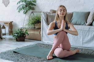 Young sporty woman holding hands in namaste gesture and practicing Ardha Matsyendrasana asana with closed eyes during morning meditation at home in living room, wellbeing and healthy life concept