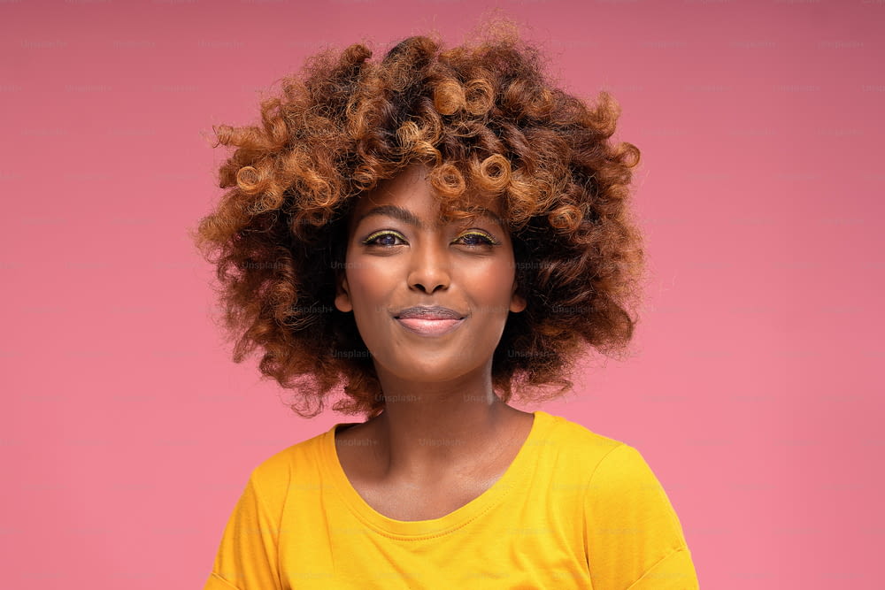 Feliz menina afro bonita com maquiagem de verão glamour e cabelo encaracolado sorrindo para a câmera no fundo de estúdio pastel rosa. Foto colorida. Emoções de pessoas reais.