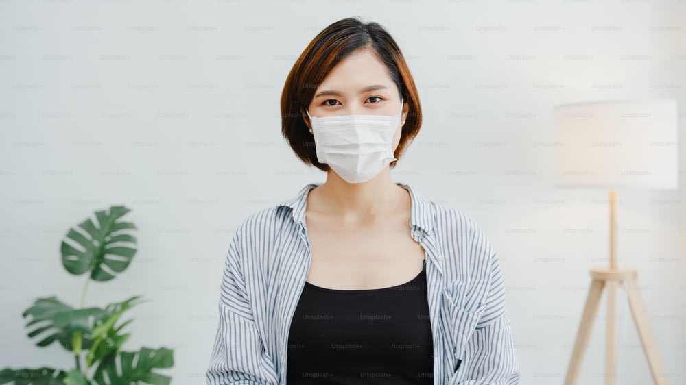Asia businesswoman wear face mask social distancing in situation for virus prevention looking at camera smiling under surgical mask back to work in office workspace. Lifestyle after corona virus.