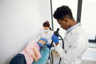 Primer plano de un médico afroamericano masculino con un endoscopio y una enfermera colega examinando a una paciente joven. Endoscopia, examen médico