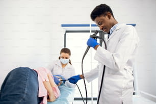 Joven médico afroamericano y enfermera que hace el procedimiento de endoscopia para el paciente femenino, acostado en el sofá en la sala de examen moderna en la clínica