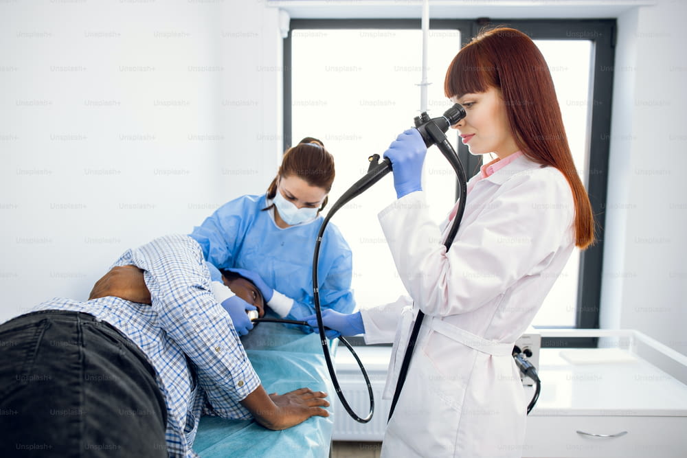 Gastroenteróloga médica concentrada femenina, endoscopio operativo durante el procedimiento gastroscópico de un joven paciente afroamericano, acostado en el sofá