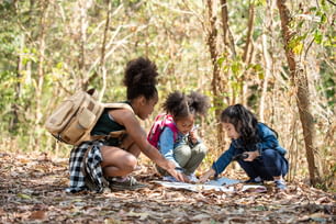 Grupo de amigas de la diversidad con mochila caminando juntas en la montaña del bosque en un día soleado de verano. Tres niños divirtiéndose en actividades al aire libre sentados y mirando el mapa explorando el bosque.