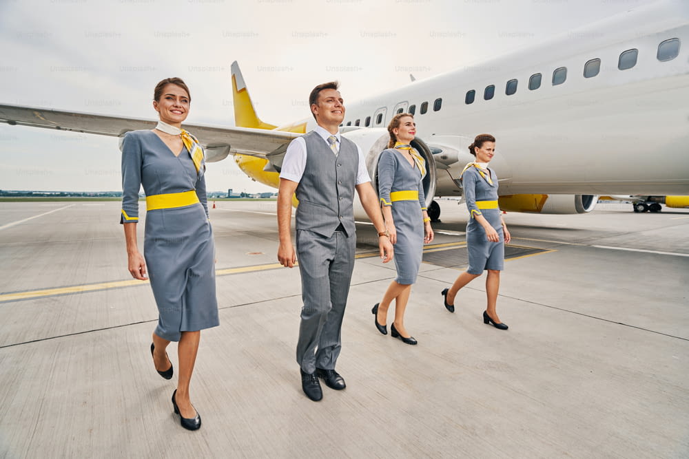 Ganzkörperporträt eines lächelnden Stewards und drei Stewardessen in Uniformen, die über den Laufsteg gehen