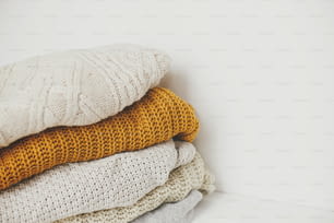 Pile of white, yellow and beige knit sweaters close up in white scandinavian room. Cozy knitted sweaters. Hello autumn. Stylish wardrobe for cold season. Space for text