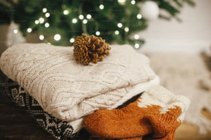 Comodi maglioni lavorati a maglia e calzini di lana su legno rustico con pigna sullo sfondo delle luci dell'albero di Natale. Mucchio di elegante stoffa invernale in una stanza scandinava decorata festosamente. Spazio per il testo