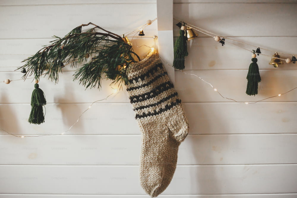 전나무 가지가 있는 크리스마스 소박한 스타킹과 크리스마스 조명이 있는 흰색 나무 벽에 종과 나무가 있는 세련된 화환. 축제 장식된 분위기의 스칸디나비아 객실. 현대적인 수제 장식