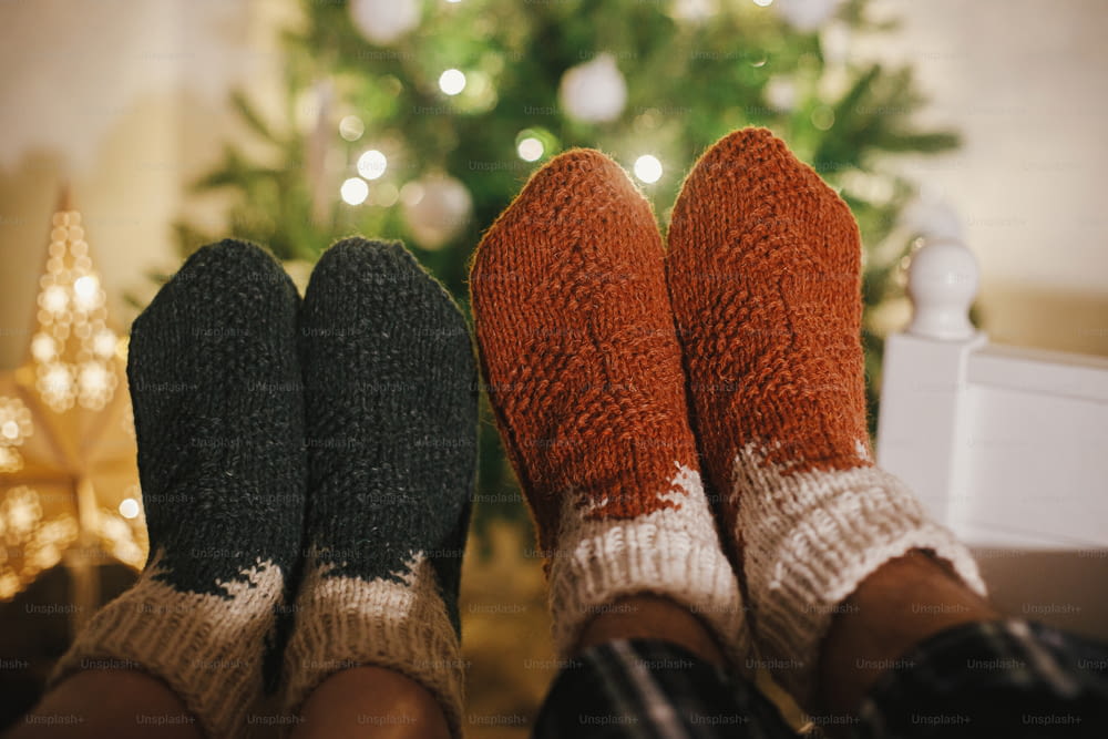 Pies de parejas en calcetines de lana acogedores en el fondo del árbol de Navidad en luces en la sala de noche festiva. Celebrando juntos las vacaciones de invierno, acogedores momentos familiares en casa. Calcetines cálidos y elegantes
