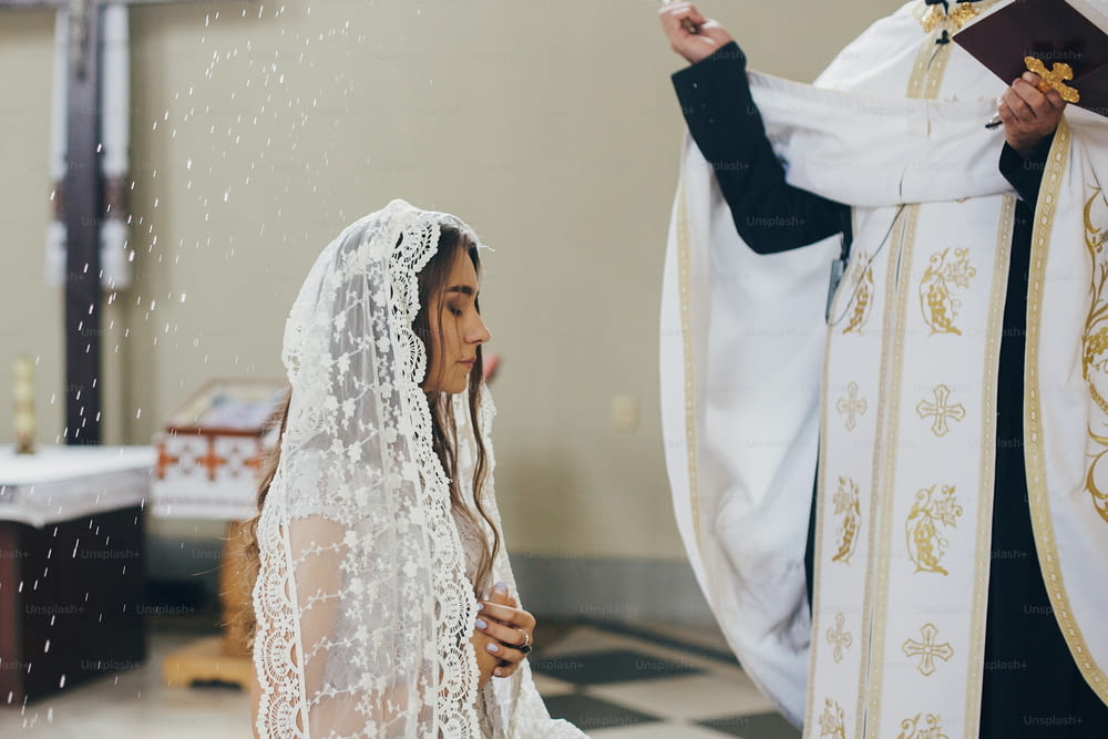 Bênção do sacerdote com água benta noiva elegante em lenço no altar durante o santo matrimônio na igreja. Cerimônia de casamento na catedral. Noiva espiritual clássica do casamento rezando