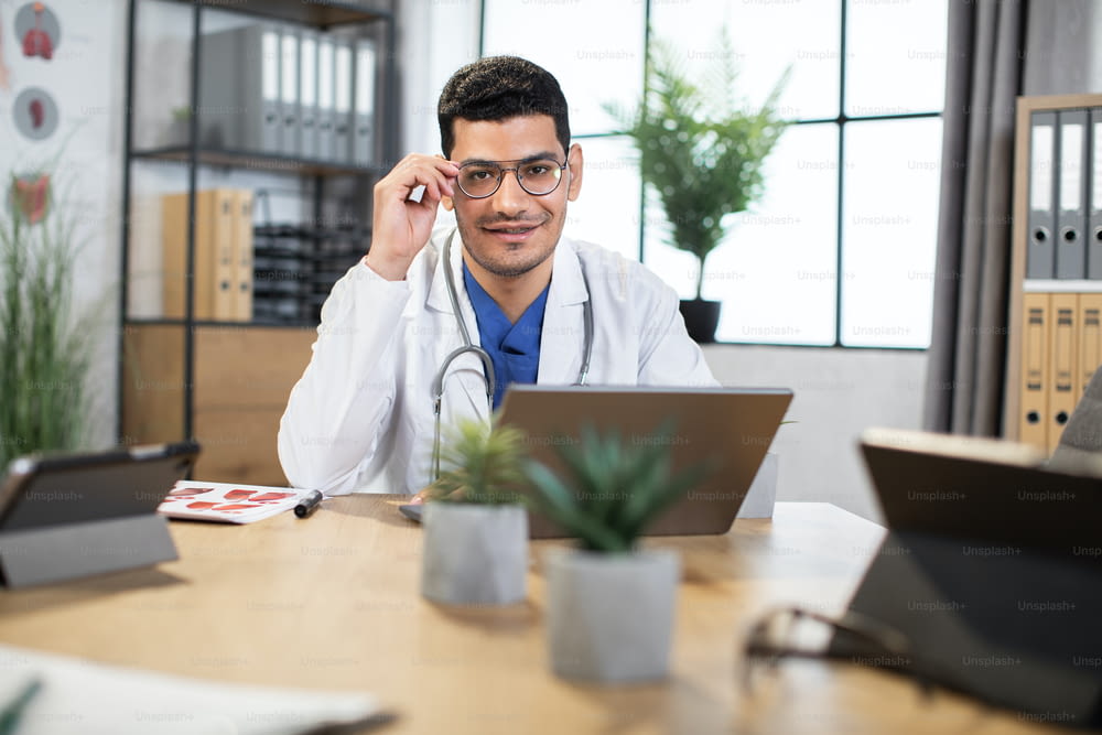 Male Arab doctor or medical student, sitting at the table indoors in modern office, and working with laptop. Young handsome male Indian physician using laptop at work, adjusting his eyeglasses