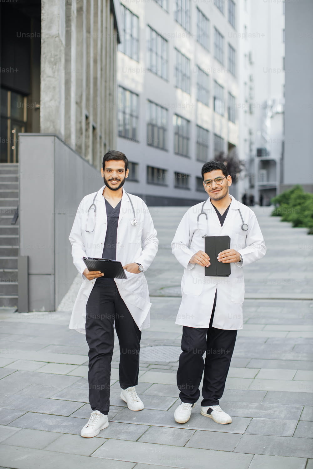 Team of two young handsome Saudi Arabian male doctors smiling outside hospital, walking with tablet pc and clipboard in hands. Full length portrait of two male medical students in white coat outdoors
