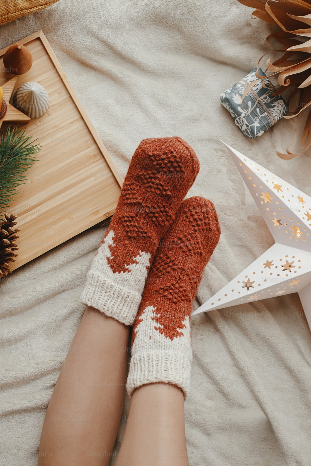 Woman legs in warm socks on soft blanket with christmas star, trees and candle, top view. Cozy moments at home. Winter and autumn holidays.