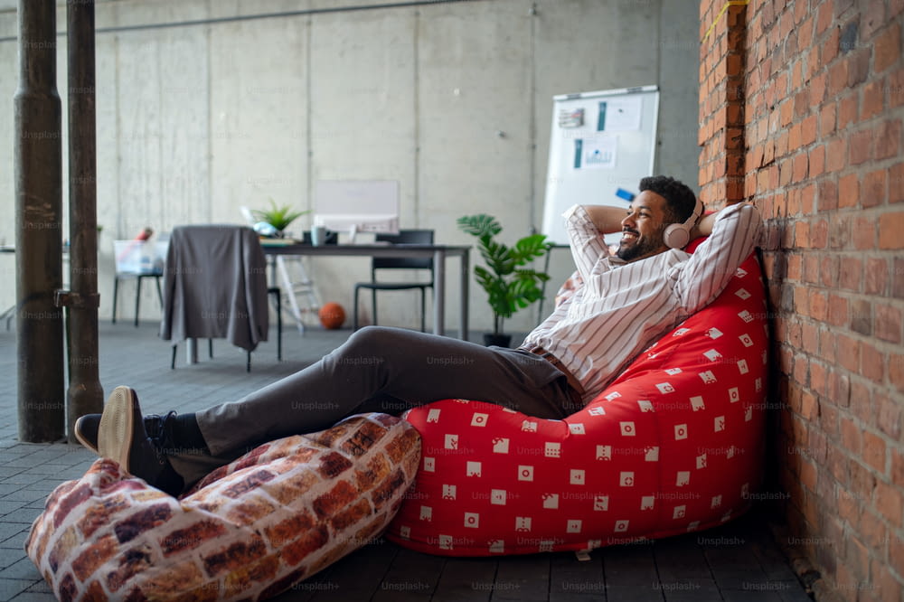 A cheerful young businessman with headphones lying on bean bag in office, taking a break and relaxing.