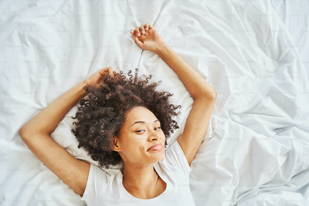 Smiling pleased relaxed beautiful young curly-haired female lying on her back on the crumpled bedsheet