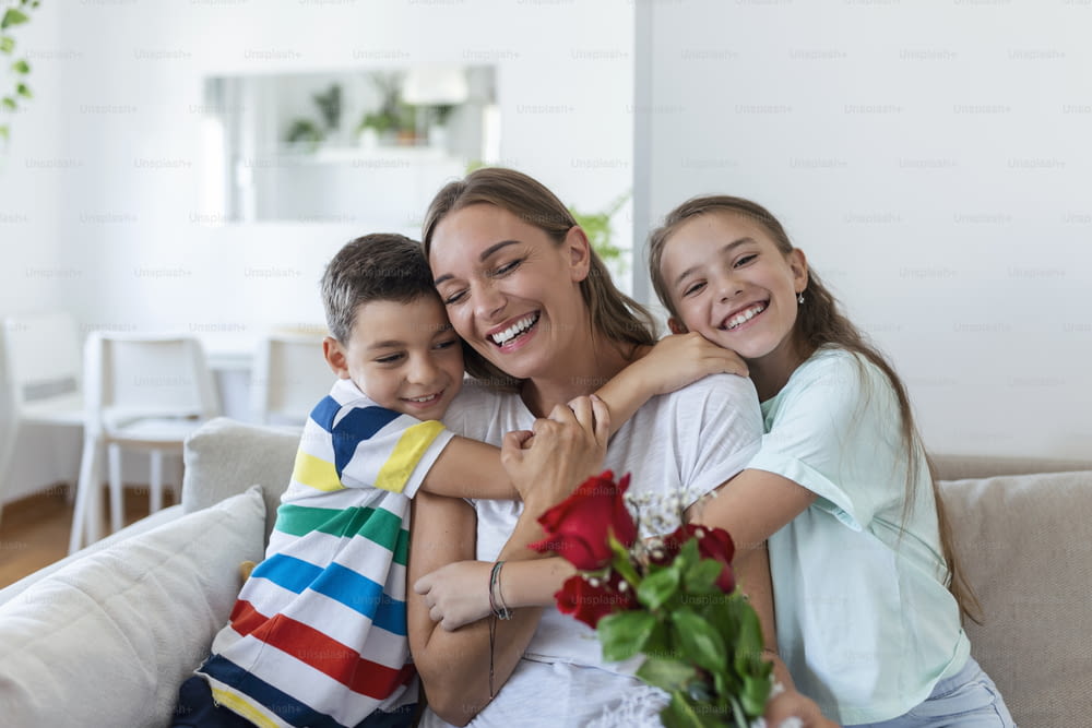 Young mother with a bouquet of roses laughs, hugging her son, and ?heerful girl with a card congratulates mom during holiday celebration in kitchen at home