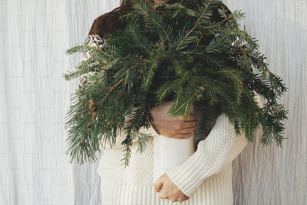 Woman in cozy sweater holding pine and fir branches in vase in rustic room. Preparations for winter holidays. Atmospheric winter time. Scandinavian hygge