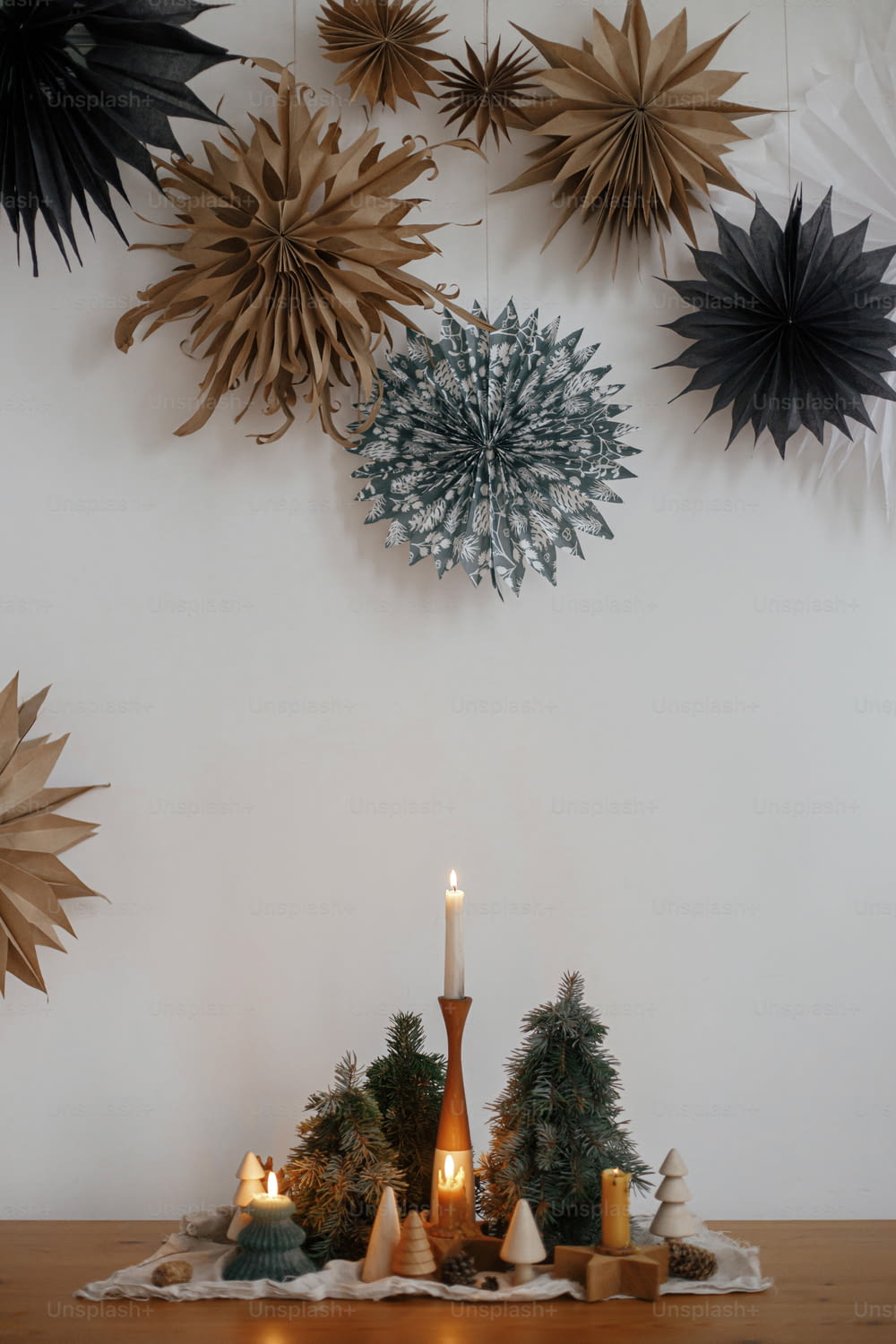 Stylish christmas candles and trees decorations on wooden table on background of white wall with big paper stars. Handmade holiday decor. Atmospheric winter time. Merry Christmas!