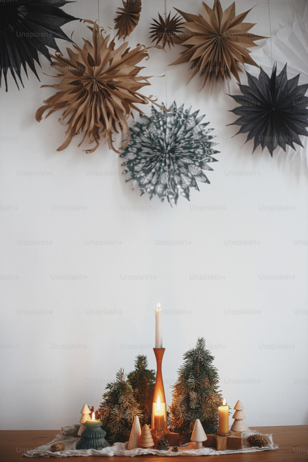 Stilvolle Weihnachtskerzen und Baumdekorationen auf Holztisch auf dem Hintergrund der weißen Wand mit großen Papiersternen. Handgemachtes Urlaubsdekor. Stimmungsvolle Winterzeit. Frohe Weihnachten!