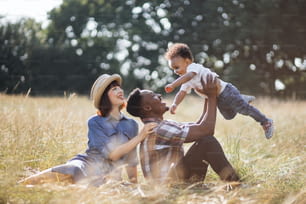 Famille multiraciale de trois personnes qui passe l’été à pique-à l’air frais. Père africain jouant avec son petit fils tandis que la mère caucasienne est assise près et sourit sincèrement.