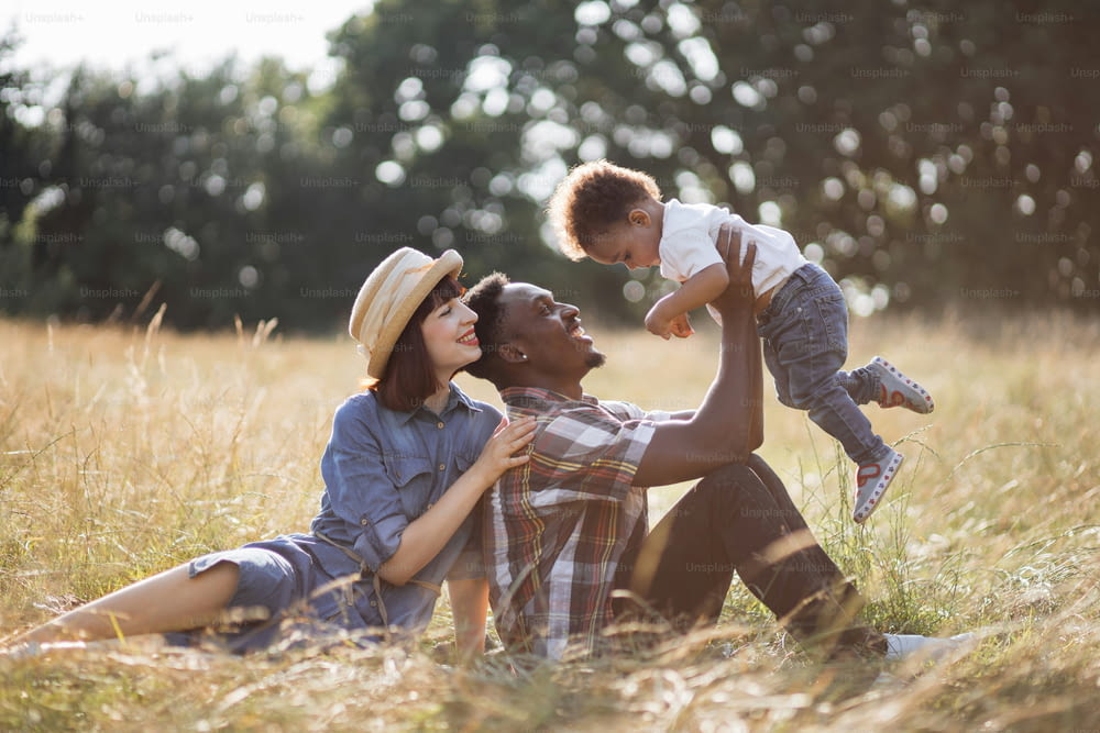 화창한 날 야외에서 함께 노는 캐주얼웨어를 입은 3명의 긍정적인 다민족 가족. 백인 여자는 아프리카 남편을 껴안고 작은 아들을 손에 들고 있습니다.