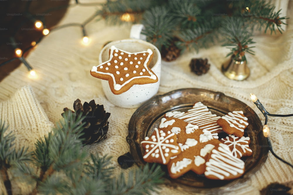 Biscotto di pan di zenzero a forma di stella di Natale su tazza di caffè calda, rami di abete, ornamenti, biscotti e luci calde su un accogliente sfondo lavorato a maglia. Tempo festivo atmosferico e casa hygge. Buone Feste