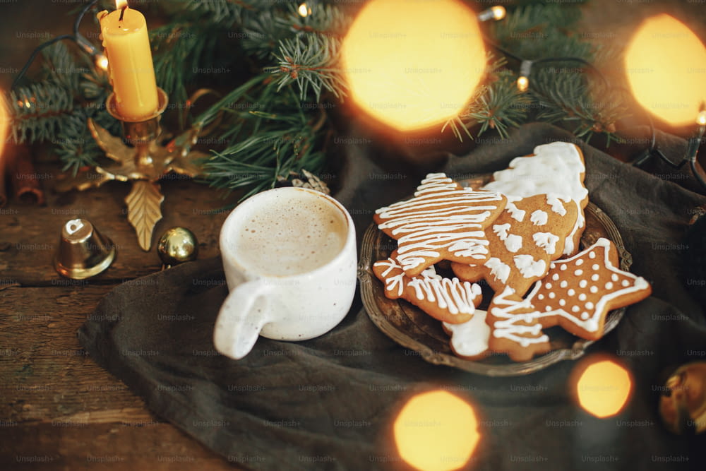 Weihnachtslebkuchenplätzchen, Kaffee in stilvoller weißer Tasse, Tannenzweige, warme Lichter auf Serviette und rustikaler Holztisch. Atmosphärisches Bild. Winter Landschaft Hygge. Frohe Feiertage!
