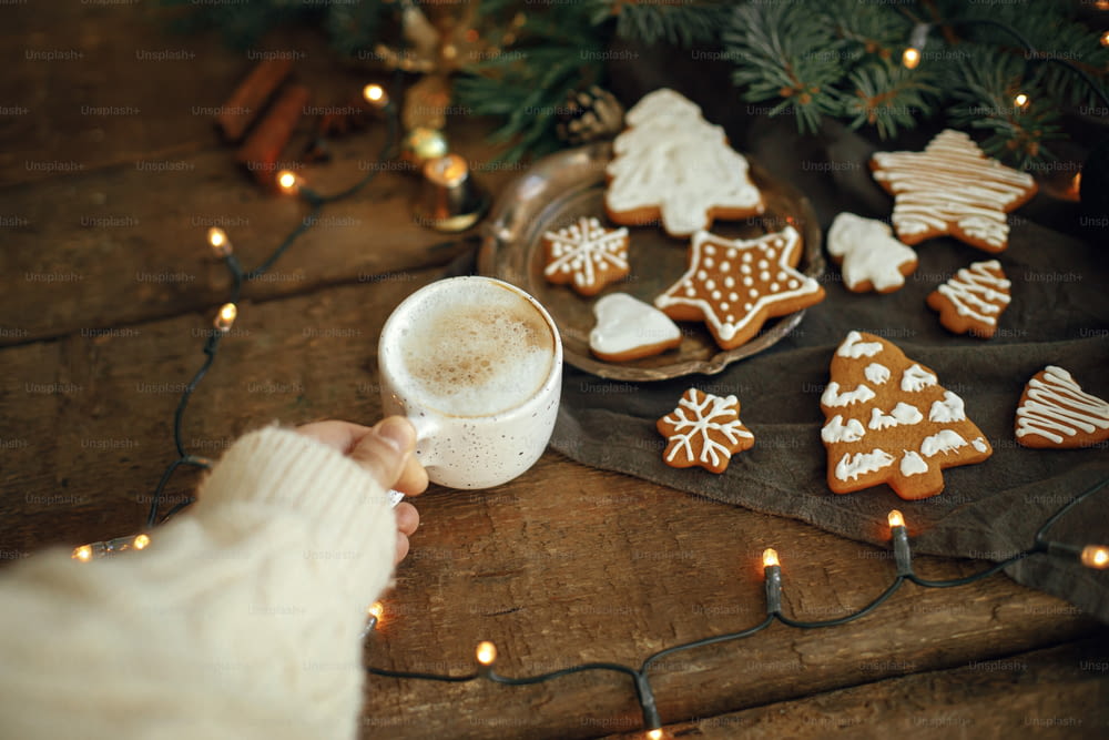 Main tenant une tasse de café chaude sur fond de biscuits en pain d’épice de Noël, de branches de sapin, de lumières chaudes sur une serviette et d’une table en bois rustique. Bonjour l’image atmosphérique de l’hiver Moody