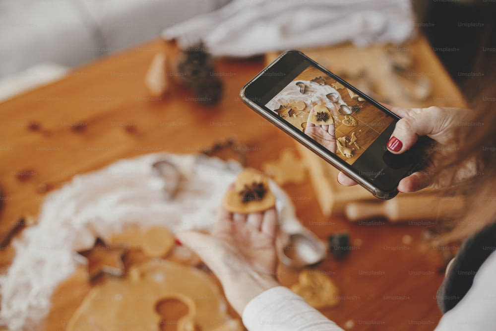 Woman holding phone and taking photo of gingerbread cookies christmas cookies on messy table flat lay. Instagram photo, social media and blog. Holiday preparations