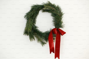 Stylish christmas wreath with red bow hanging on white wall. Merry Christmas and Happy Holidays! Simple traditional xmas wreath with pine branches and red ribbon isolated on white