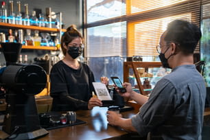 L'uomo asiatico indossa una maschera protettiva per il viso durante la pandemia di COVID-19 utilizzando il codice a barre di scansione dello smartphone effettua il pagamento contactless al bar. Piccole imprese con il concetto di pagamento elettronico mobile banking