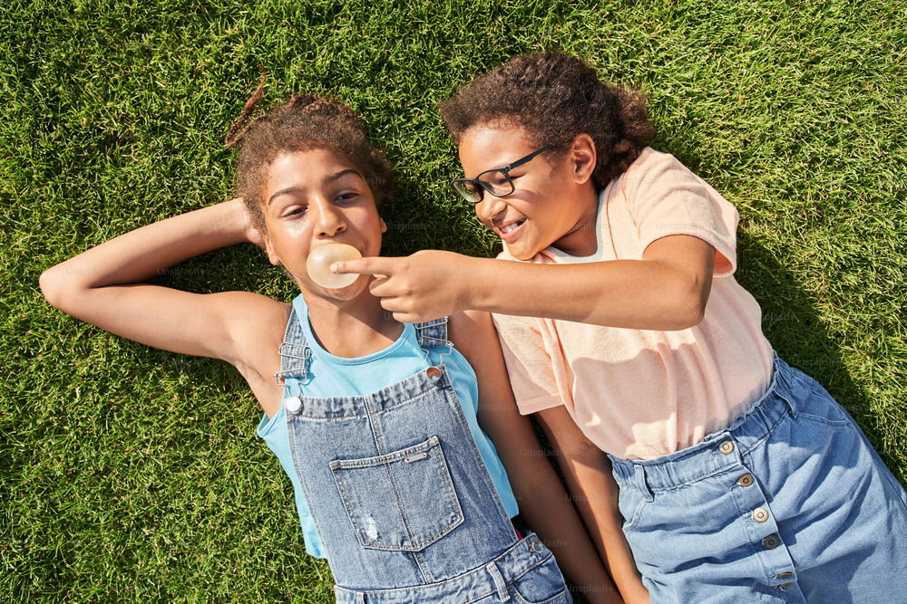 Little girl breaking chewing gum of her girlfriend outdoor. Concept of childhood. Idea of friendship. Modern kid lifestyle. Joking curly brunette girls lying on green grass.