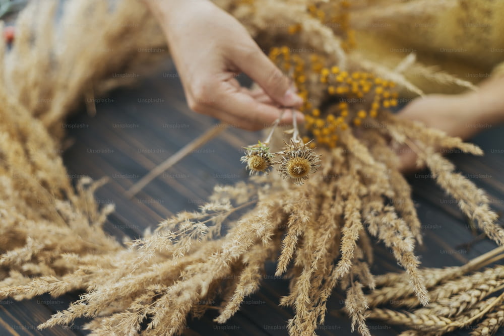 Making stylish autumn boho wreath with dry grass and wildflowers on rustic wooden table. Holiday workshop. Florist in yellow sweater making rustic wreath on dark wood