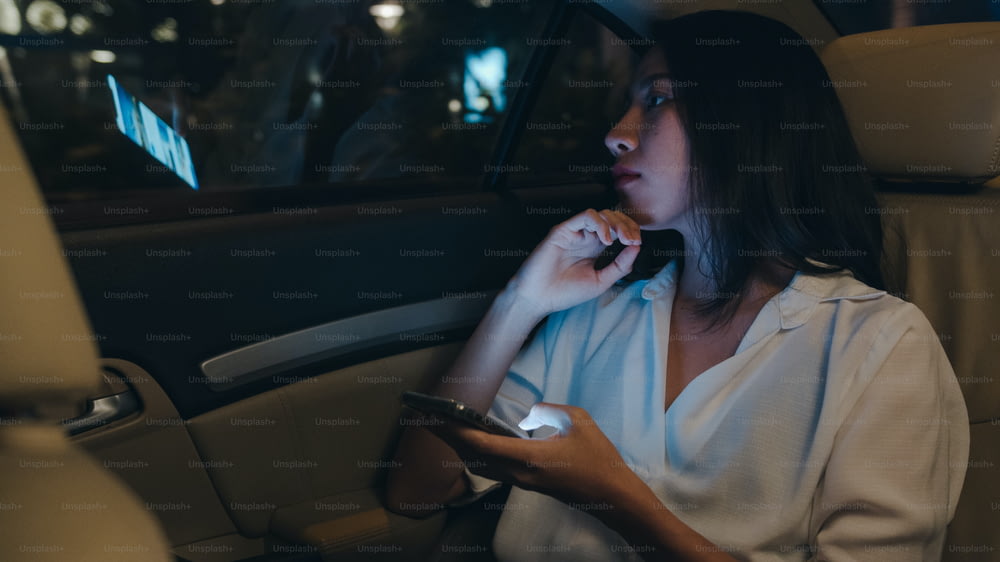 Erfolgreiche junge asiatische Geschäftsfrau in Mode-Bürokleidung arbeitet spät mit Smartphone auf dem Rücksitz des Autos in der urbanen modernen Stadt in der Nacht. Menschen berufliches Burnout-Syndrom Konzept.