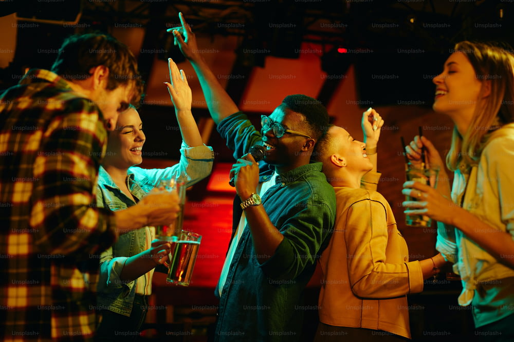 Grupo multiétnico de jovens se divertindo e cantando karaokê em um bar à noite. O foco está no homem negro.