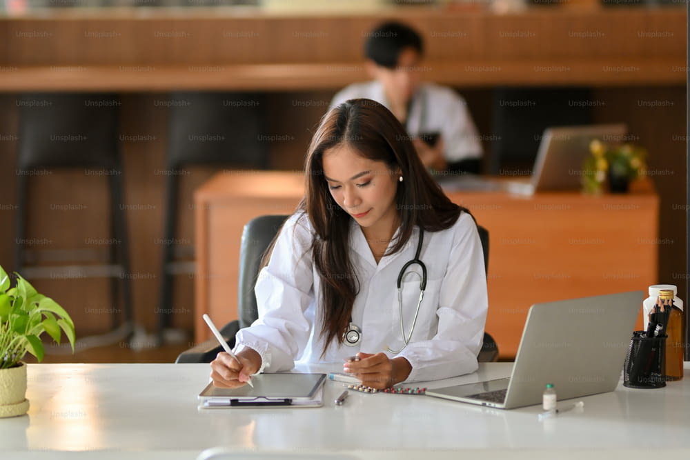 A young female doctor works on a digital tablet. In a hospital office, a physician in a white lab coat reads a past medical history research.