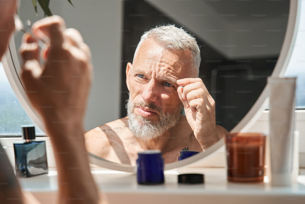 バスルームで鏡を見ながら顔に眉毛を寄せる成熟した男性。顔のスキンケアと衛生のコンセプト。家庭のライフスタイル。入れ墨と白髪のヨーロッパの男性年金受給者