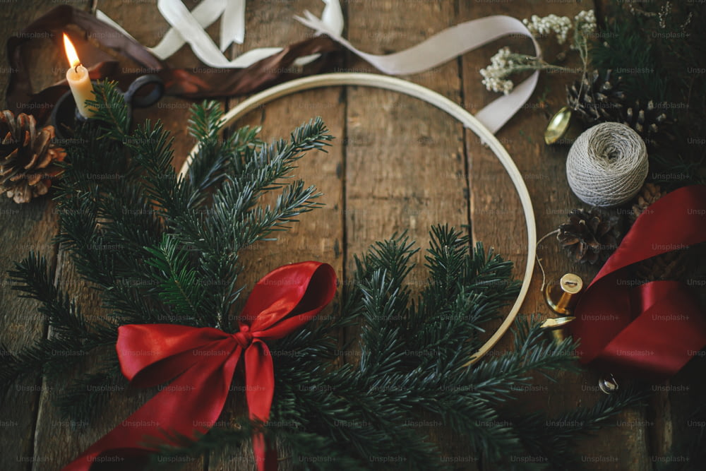 Feliz Natal e Boas Festas! Coroa de Natal moderna com ramos de abeto e laço vermelho na mesa de madeira rústica com vela, fitas, pinhas. Imagem atmosférica mal-humorada.