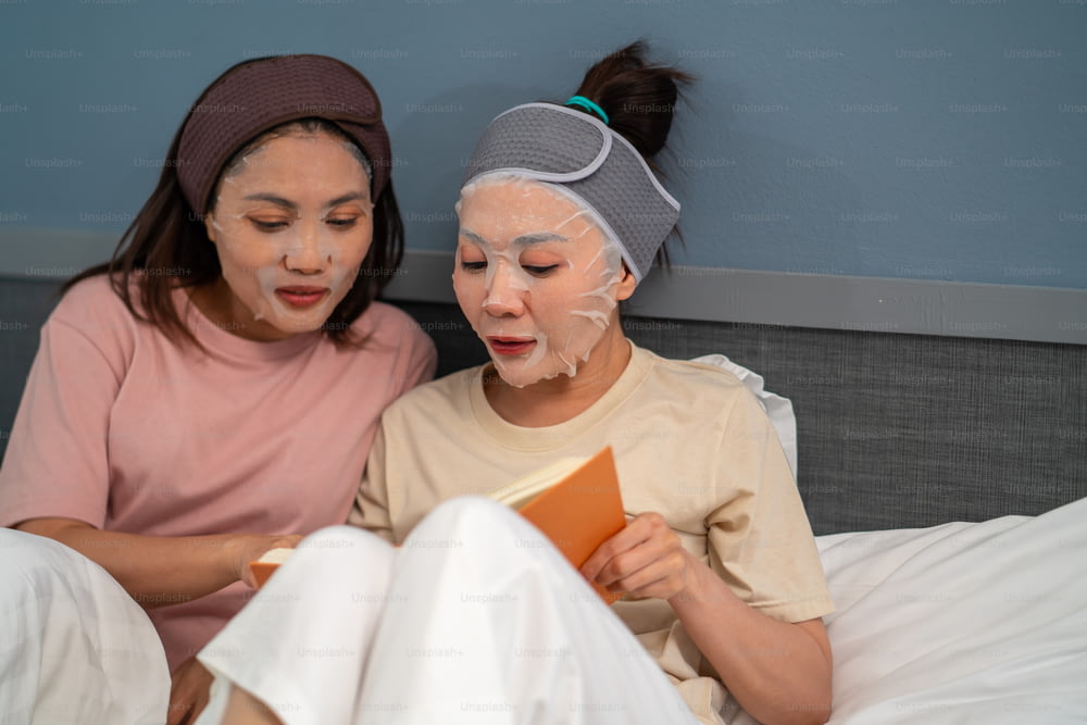 Smiling Asian woman friends sitting on the bed with applying skin care facial mask on their face together at home. Female gay couple relax and enjoy beauty facial treatment and reading a book together.