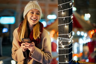 幸せな美しい若い女性の女の子は、お祝いのクリスマスフェアの背景を持つ街でスマートフォンを使用しています