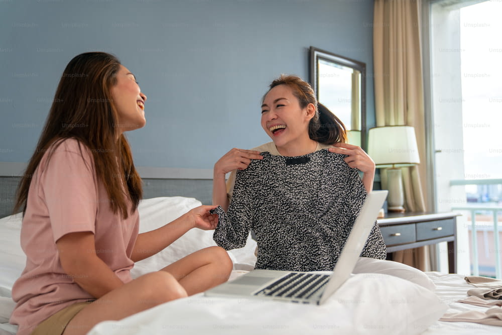 Asian woman sitting on the bed in bedroom and try on outdoor dress showing to her female friend with smiling. Female gay couple choosing clothing together at home with happiness