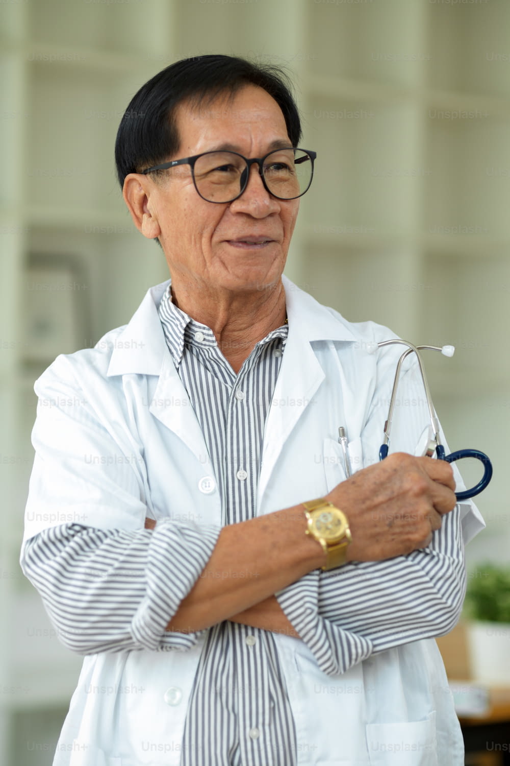 Un thérapeute asiatique d’âge moyen avec des lunettes et une blouse blanche se tient debout, les bras croisés, dans sa clinique.