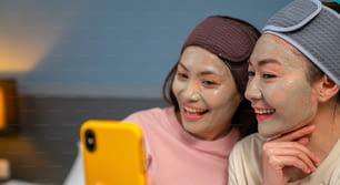 Amigas asiáticas sorridentes sentadas na cama com a aplicação de máscara facial de tratamento de cuidados com a pele em seu rosto juntas em casa. Casal gay feminino usando smartphone tirando selfie junto com a felicidade